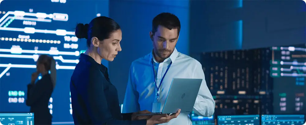 Two IT professionals looking at laptop screen in server room.